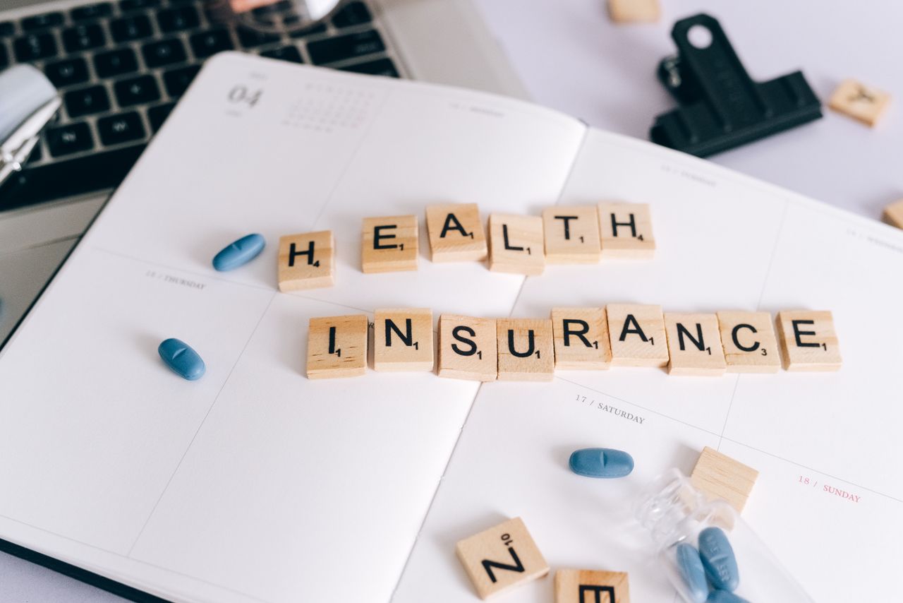 Scrabble tiles spelling Health Insurance on planner with pills and laptop, symbolizing healthcare planning.